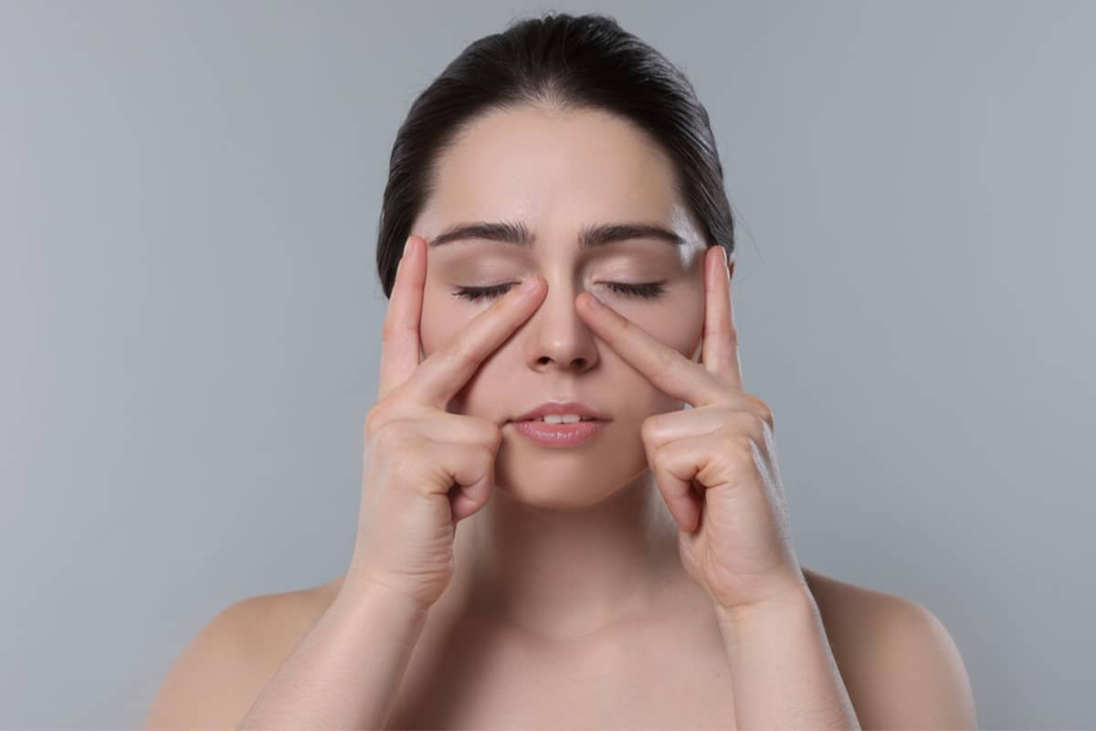 Una mujer con los ojos cerrados tocando suavemente los lados del puente nasal con los dedos para ilustrar hinchazón o sensibilidad después de un procedimiento de rinoplastia.