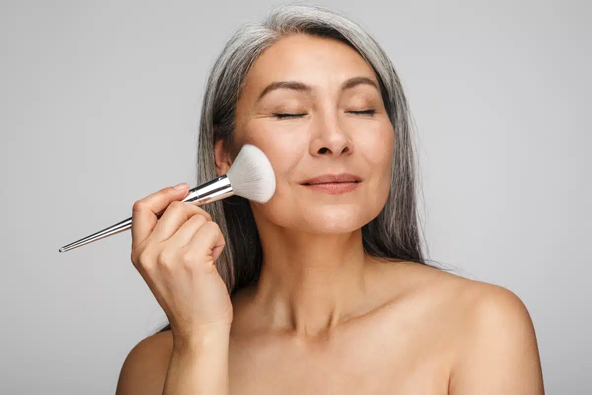 Mujer madura con cabello largo y plateado, sonriendo con los ojos cerrados mientras se aplica suavemente polvo en la mejilla con una brocha de maquillaje grande.