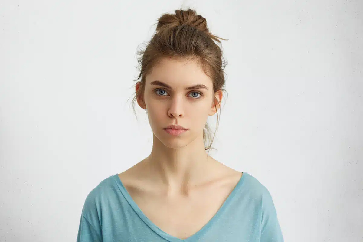 Mujer joven de tez clara y ojos azules con el cabello castaño recogido, vistiendo una camiseta azul y mirando seriamente a la cámara sobre un fondo blanco.