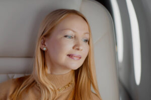 Mujer rubia con un suéter marrón claro mirando relajada por la ventana desde el asiento de un avión.