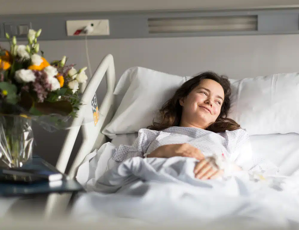 Paciente sonriente acostada en una cama de hospital con flores en la mesa auxiliar, destacando la importancia de la disponibilidad de camas hospitalarias para programar la cirugía.