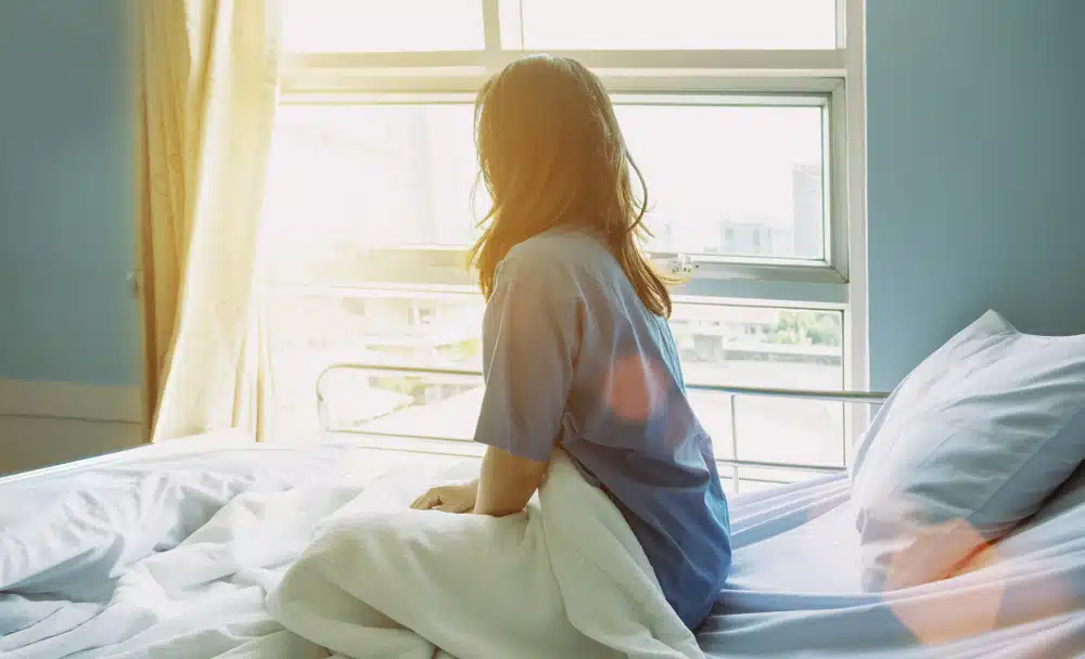 Paciente sentada en una cama de hospital mirando por la ventana con luz solar entrando, destacando el impacto de las estaciones en la recuperación quirúrgica.