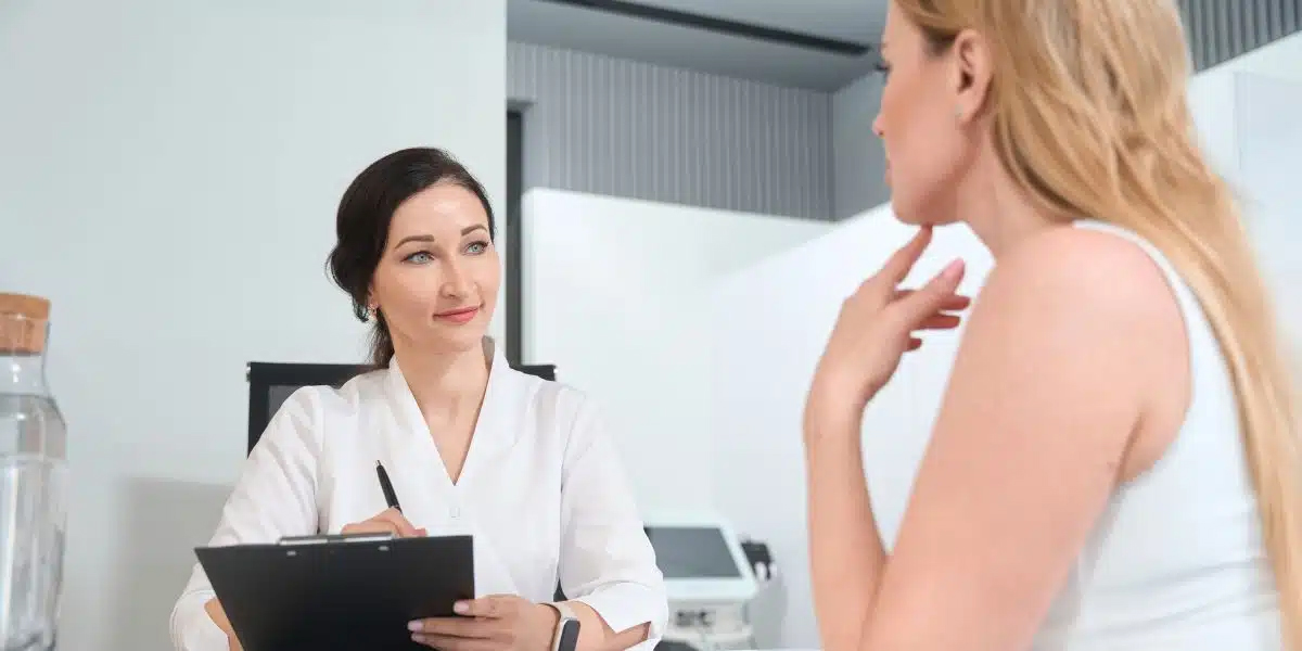 Doctora sosteniendo un portapapeles y hablando sobre los costos del lifting facial con una mujer rubia en una sala de consulta.