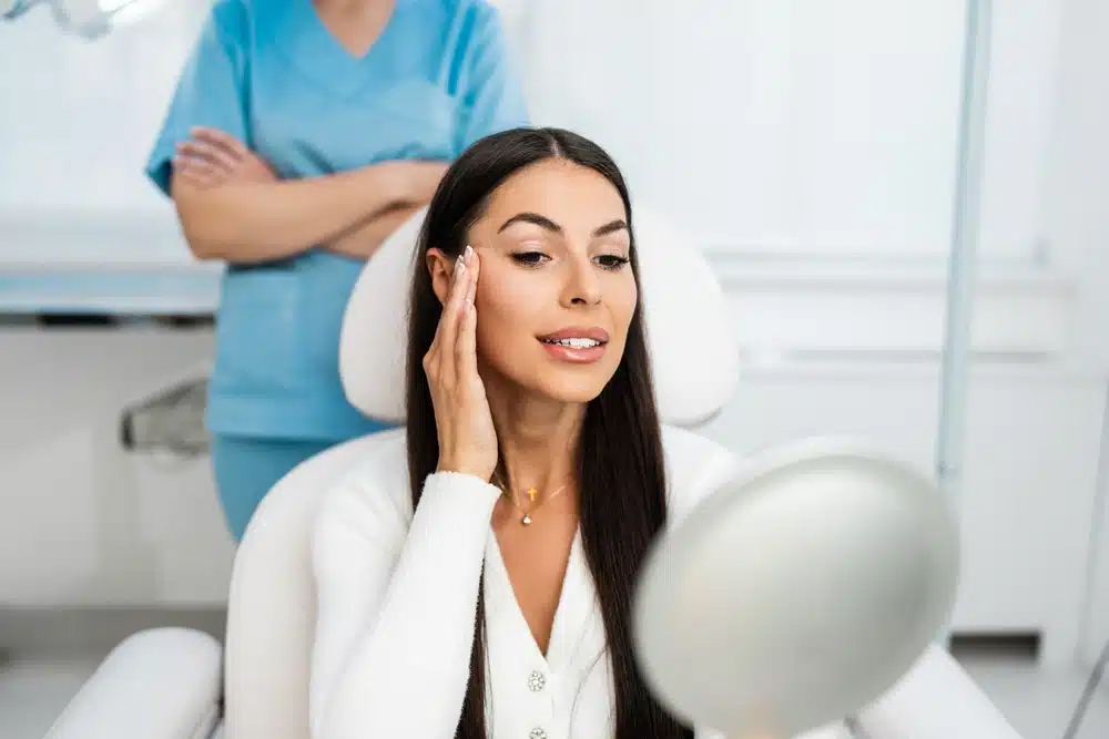 Mujer de cabello largo y oscuro mirándose el rostro en un espejo de mano, sentada en una silla médica, con un profesional médico de pie al fondo.