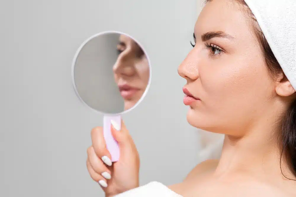 Mujer con bata y el cabello envuelto en una toalla inspeccionando su nariz en un espejo de mano después de una rinoplastia.