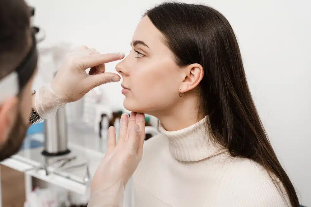 Paciente mujer con cabello oscuro en una consulta médica, con un doctor examinando su nariz para una posible rinoplastia secundaria.