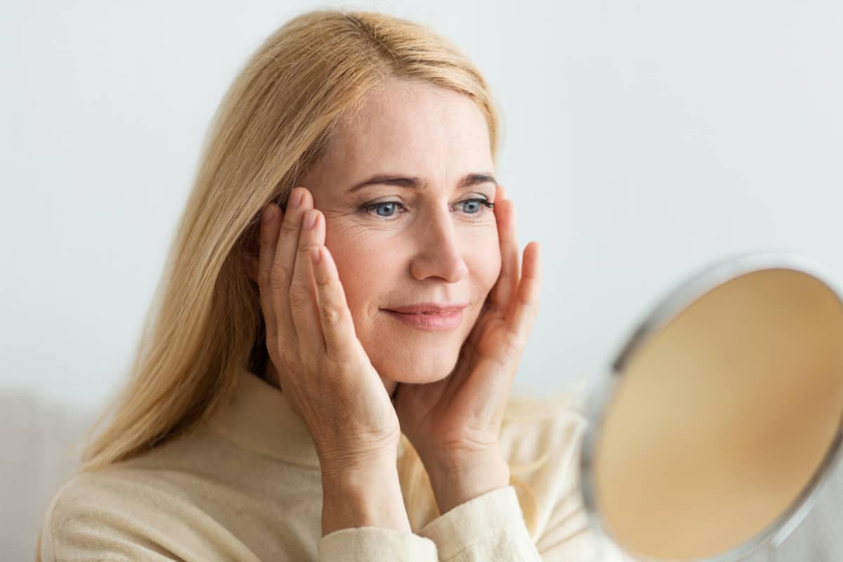 Una mujer mirándose al espejo y tocándose las sienes para controlar la maduración y atenuación de sus cicatrices de lifting facial durante la fase de recuperación.