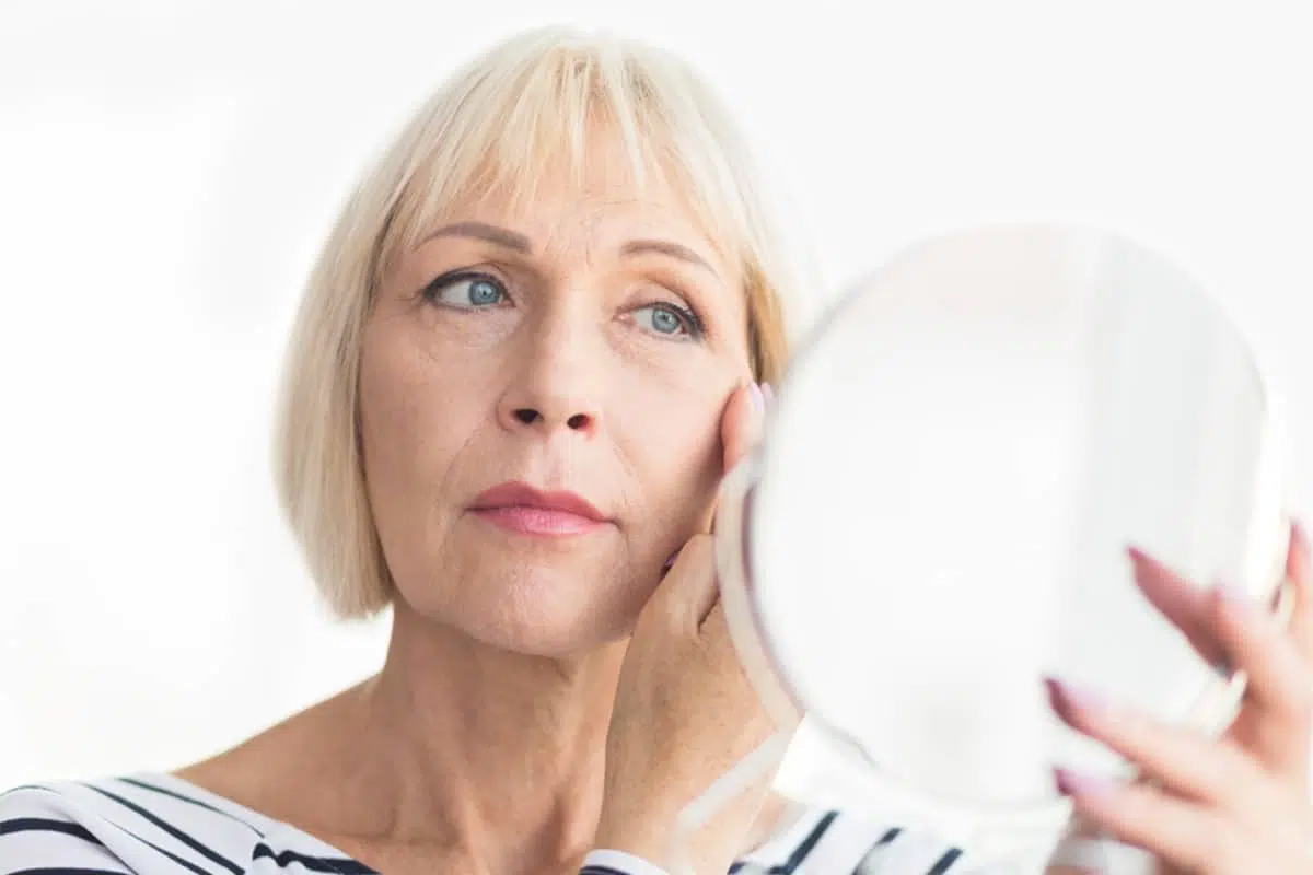 Una mujer rubia se mira en un espejo de mano para evaluar sus contornos faciales y la resolución gradual de la hinchazón después de un procedimiento de lifting facial.
