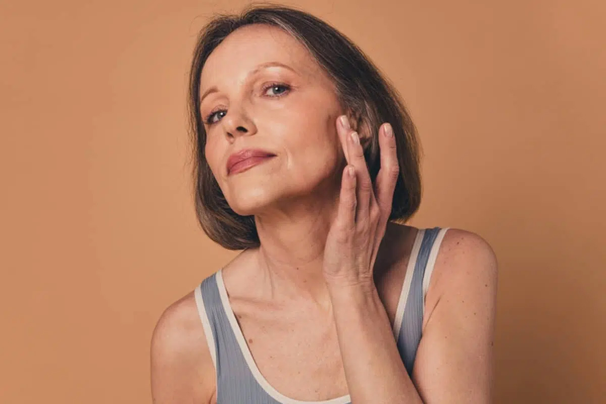 Una mujer tocándose suavemente la mejilla y la línea mandibular mientras mira hacia arriba, evaluando la elasticidad de su piel antes de someterse a un lifting facial.