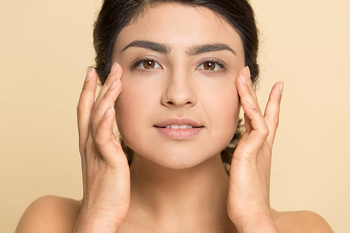 Retrato en primer plano de una mujer con cabello oscuro y ojos marrones colocando suavemente las puntas de los dedos sobre sus mejillas contra un fondo beige.