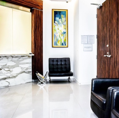 Área de recepción con modernas sillas negras, mostrador de mármol, detalles en madera y obras de arte llamativas en un consultorio médico.