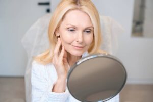 Mujer en bata blanca evaluando los contornos de su rostro en un espejo a los tres días de un lifting facial.