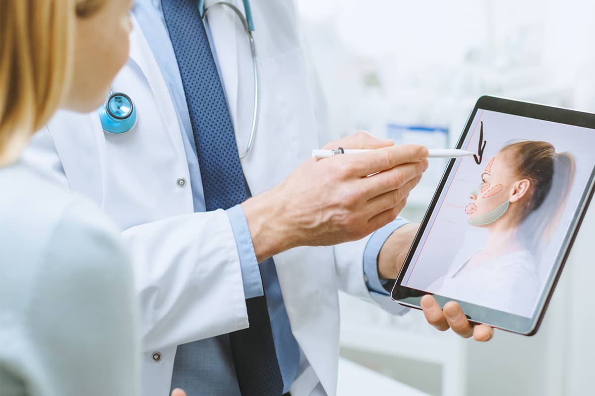 Un médico con bata blanca usa un lápiz óptico para señalar un mapa simulado de lifting facial en la pantalla de una tableta durante una consulta con una paciente.