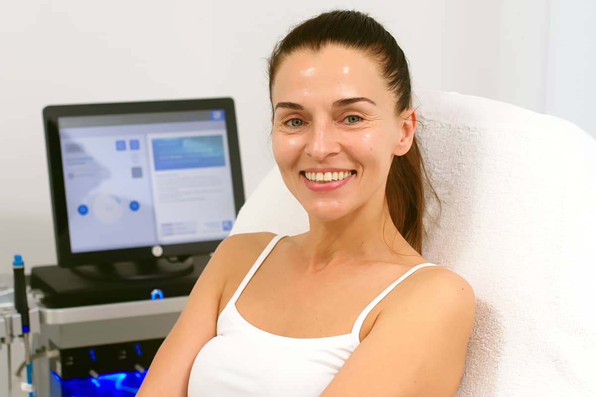 Una mujer sonriente con el cabello recogido en una cola de caballo está sentada en una silla clínica de tratamiento, con un monitor de imágenes médicas visible al fondo.