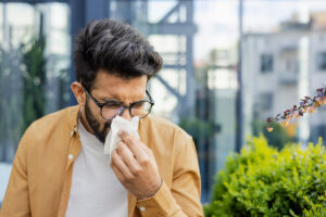Hombre joven con barba y gafas en exteriores, sonándose la nariz cuidadosamente con un pañuelo de papel blanco.