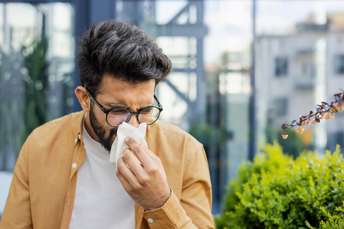 Hombre joven con barba y gafas en exteriores, sonándose la nariz cuidadosamente con un pañuelo de papel blanco.