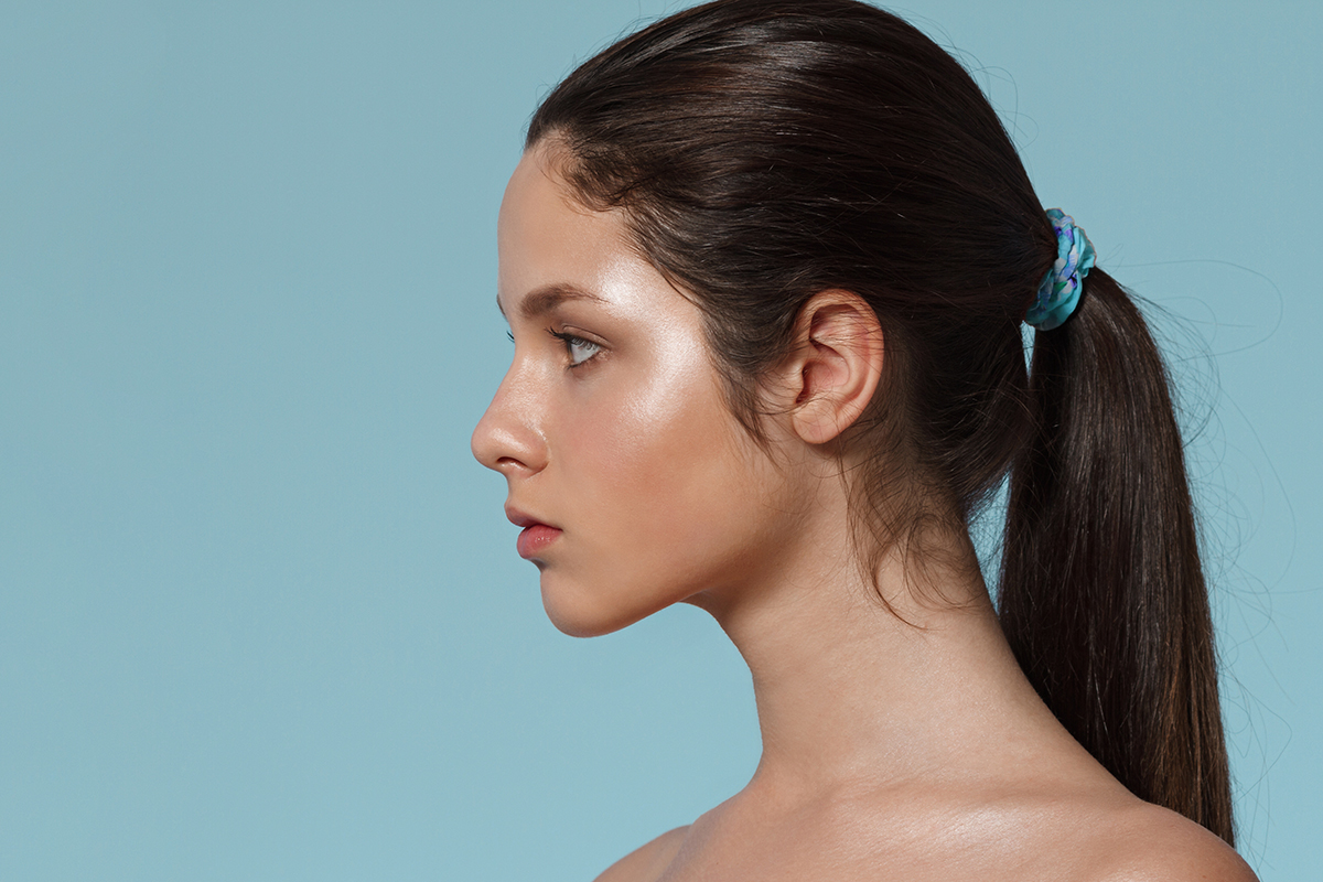 Perfil del rostro de una mujer joven con el cabello oscuro recogido en una coleta, mostrando una estructura nasal equilibrada sobre un fondo azul claro.