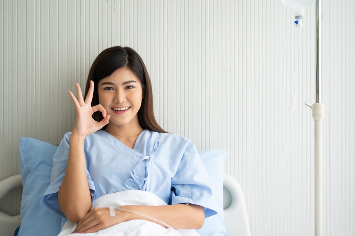 Mujer joven sonriente sentada en una cama de clínica con una bata hospitalaria azul, haciendo el gesto de 'OK' con su mano derecha.
