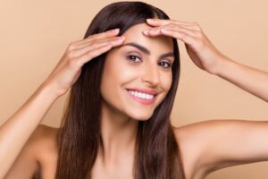 Mujer joven sonriente tocando la piel tersa de su frente, mostrando los posibles resultados de un lifting de cejas.