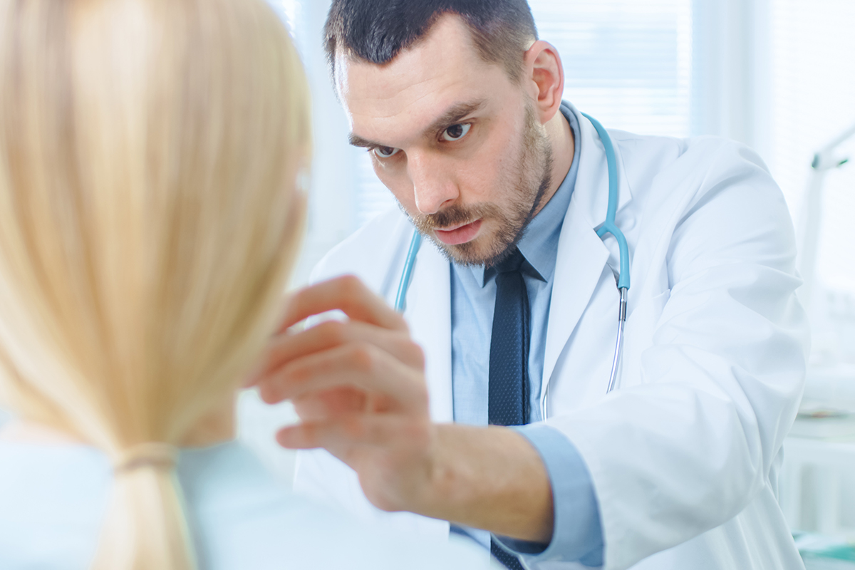 Médico especialista con bata blanca examinando cuidadosamente el rostro y cuello de una paciente rubia en su consultorio.