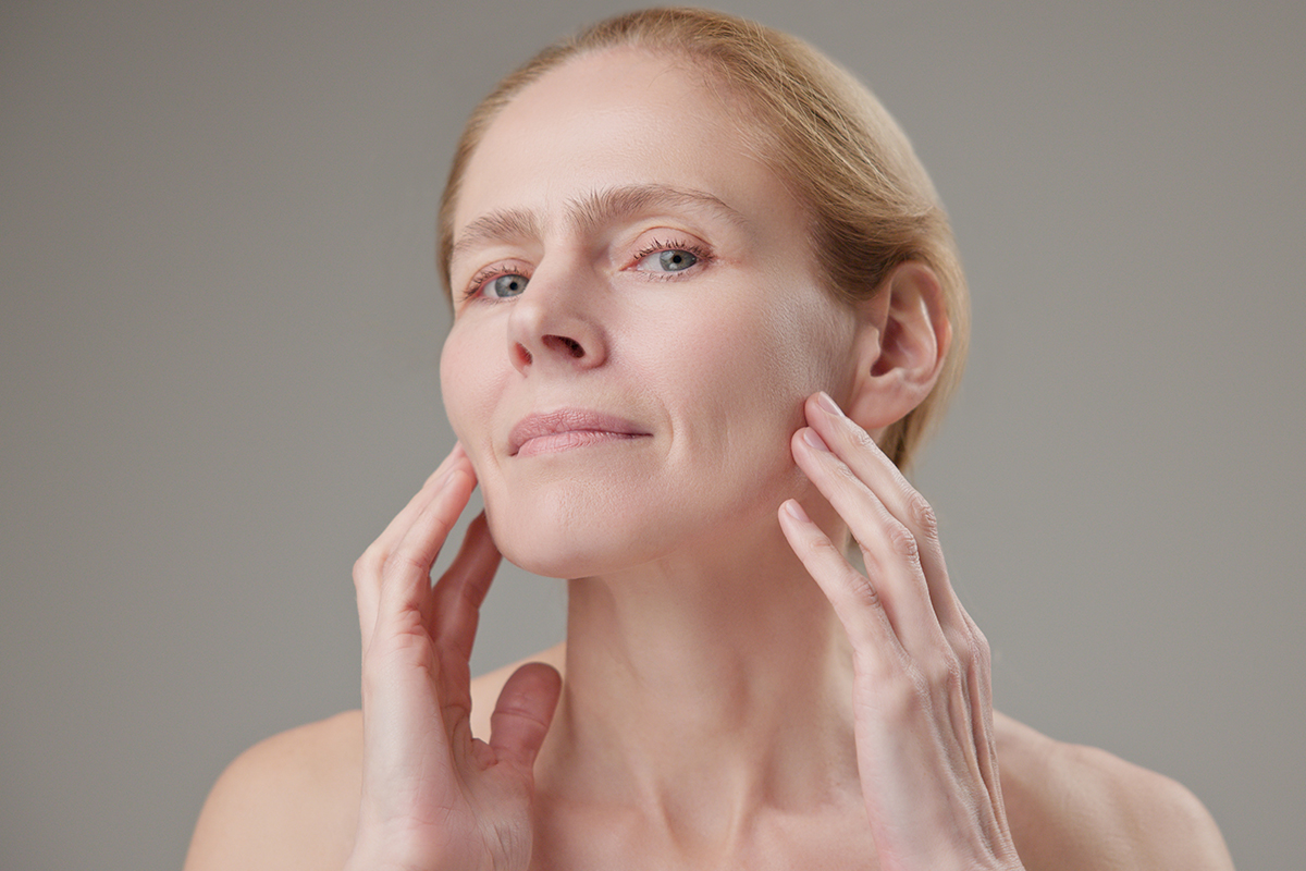 Una mujer toca suavemente la zona media del rostro y la mejilla para evaluar la firmeza y el grado de hinchazón después de un lifting facial de plano profundo.