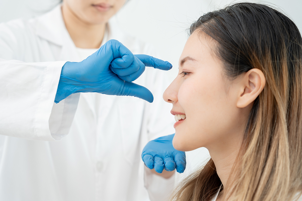 Especialista con guantes azules evaluando el perfil nasal de una paciente sonriente durante una consulta de rinoplastia.