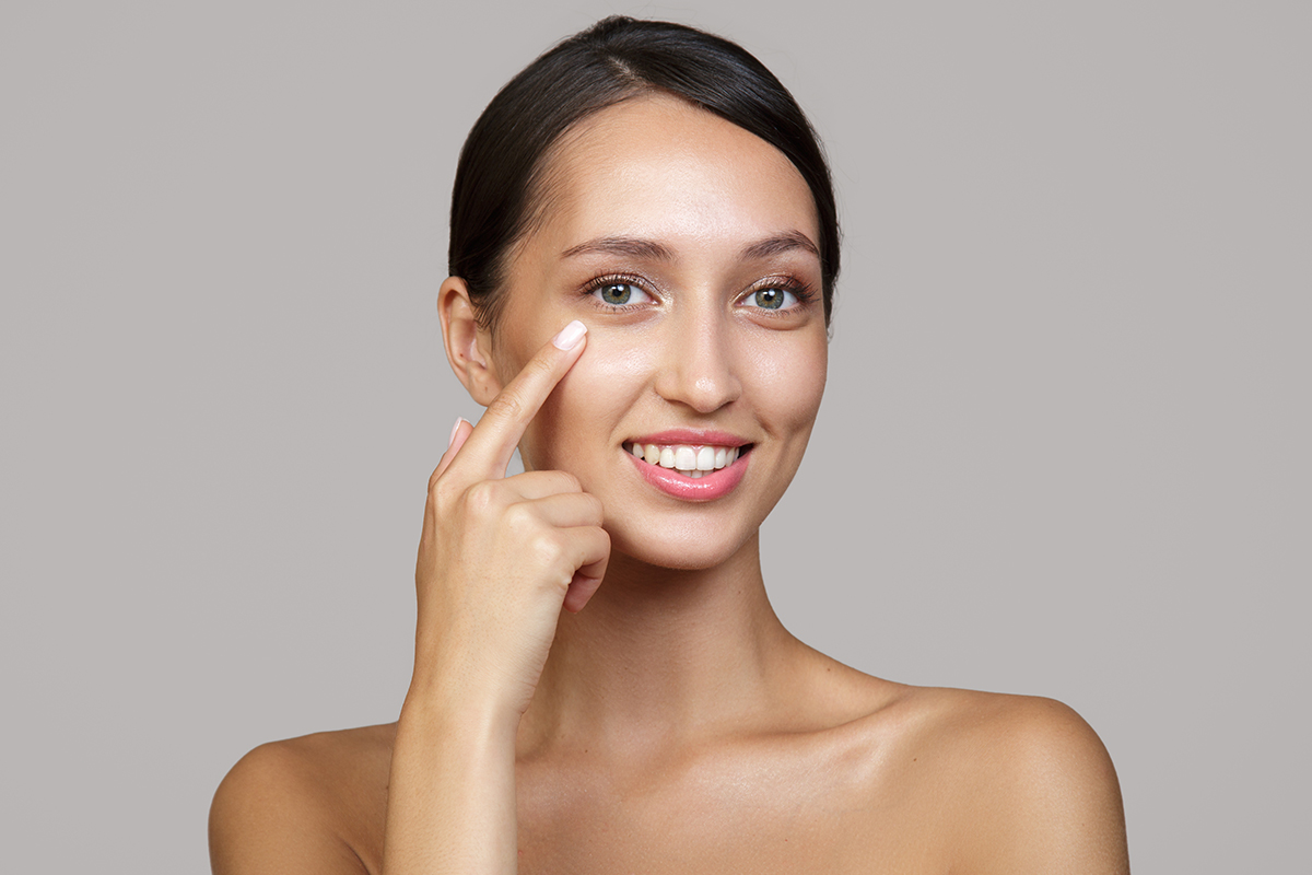 Mujer joven sonriendo y señalando suavemente la zona debajo de su ojo, mostrando una piel impecable.