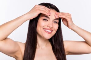 Mujer joven y sonriente tocando su frente y cejas con ambas manos, mostrando un rostro rejuvenecido.