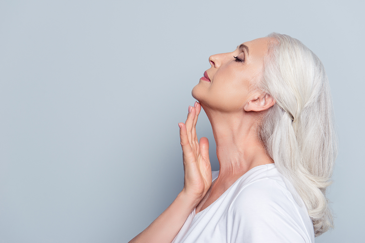 Mujer madura de perfil con cabello plateado, tocando suavemente la piel firme y tersa de su cuello y línea de la mandíbula.