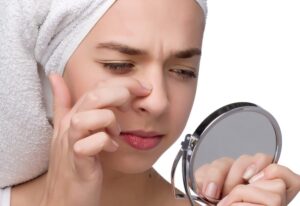 Mujer examinando su nariz hinchada frente al espejo tras una operación de rinoplastia, con toalla blanca en la cabeza.