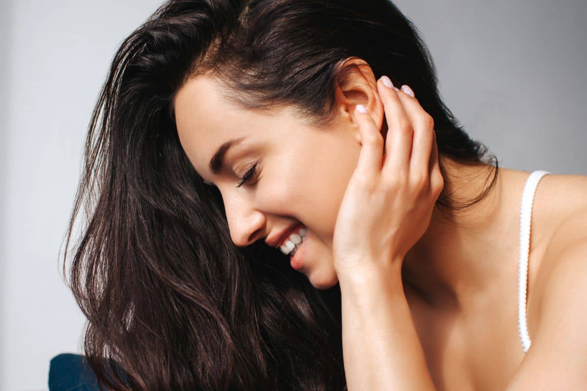 Mujer sonriendo con la cabeza inclinada y la mano cerca de la oreja, mostrando confianza tras una otoplastia.