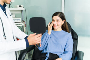 Mujer joven sonriendo feliz y mirándose en un espejo de mano sostenido por un médico, tocando suavemente el vendaje de su nariz en el consultorio.