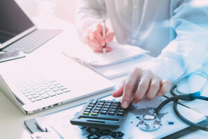 Manos de un profesional médico usando una calculadora y escribiendo en un bloc de notas sobre un escritorio con un estetoscopio y una computadora portátil.