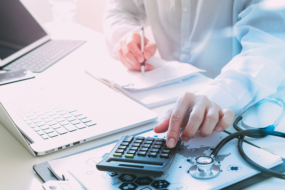 Manos de un profesional médico usando una calculadora y escribiendo en un bloc de notas sobre un escritorio con un estetoscopio y una computadora portátil.