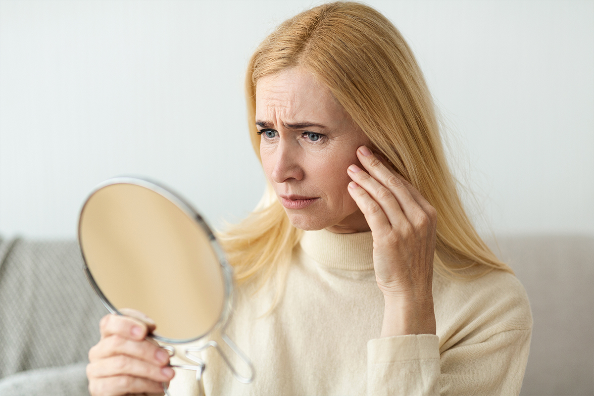 Mujer preocupada mirándose al espejo y tocando su rostro para evaluar la flacidez en cuello y mandíbula.